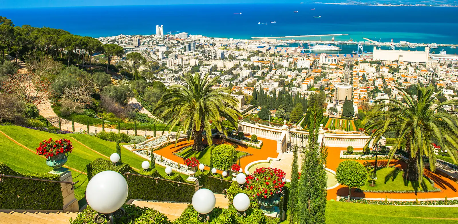 View of the stunning Bahai gardens with palm trees and flowerbeds overlooking Haifa’s coastal city and harbor, with the sea and ships in the background.