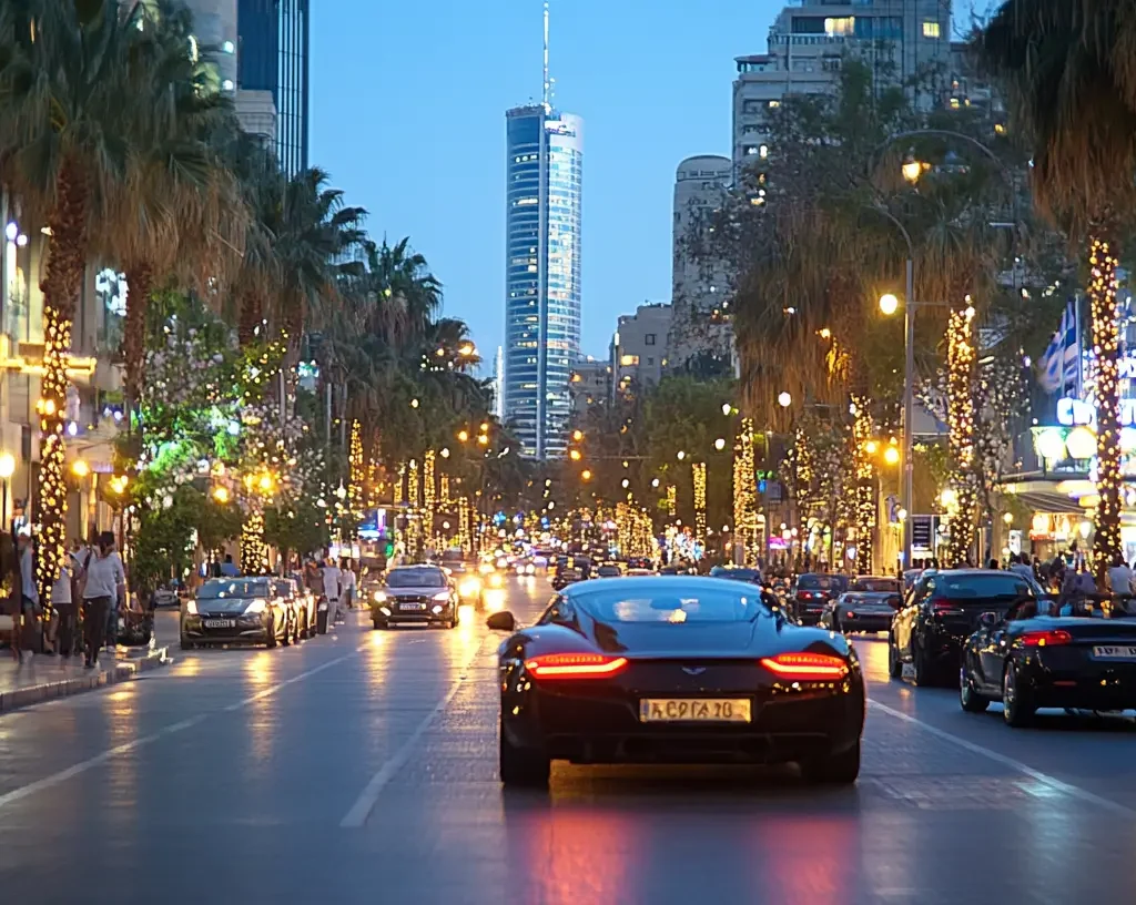 A sleek black sports car drives down a lively, palm-lined city street at dusk, surrounded by lit-up buildings, illuminated trees, and other cars, with a tall skyscraper in the background.