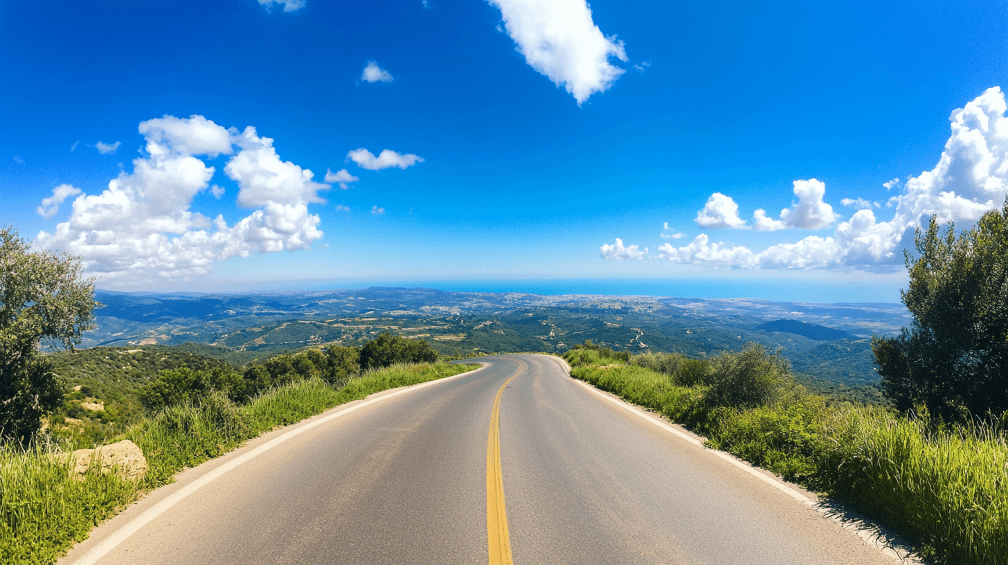 A winding road stretches downhill through lush green hills, with expansive views of valleys, forests, and distant blue sea under a bright blue sky dotted with fluffy white clouds.