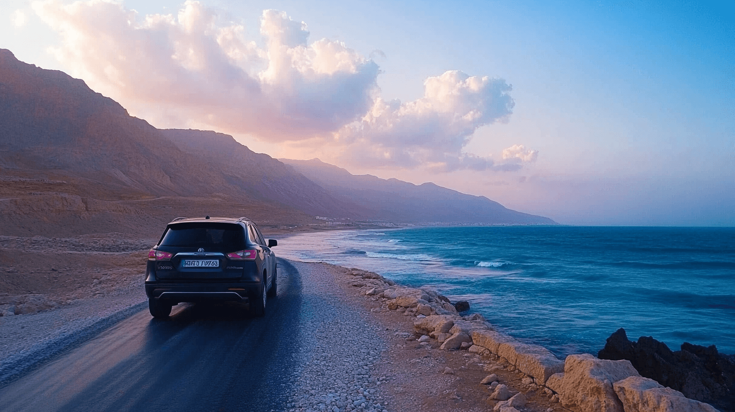 A car drives along a coastal road in Israel at sunset, with mountains on the left and the blue ocean on the right under a sky with pink clouds—a perfect tiyul moment.
