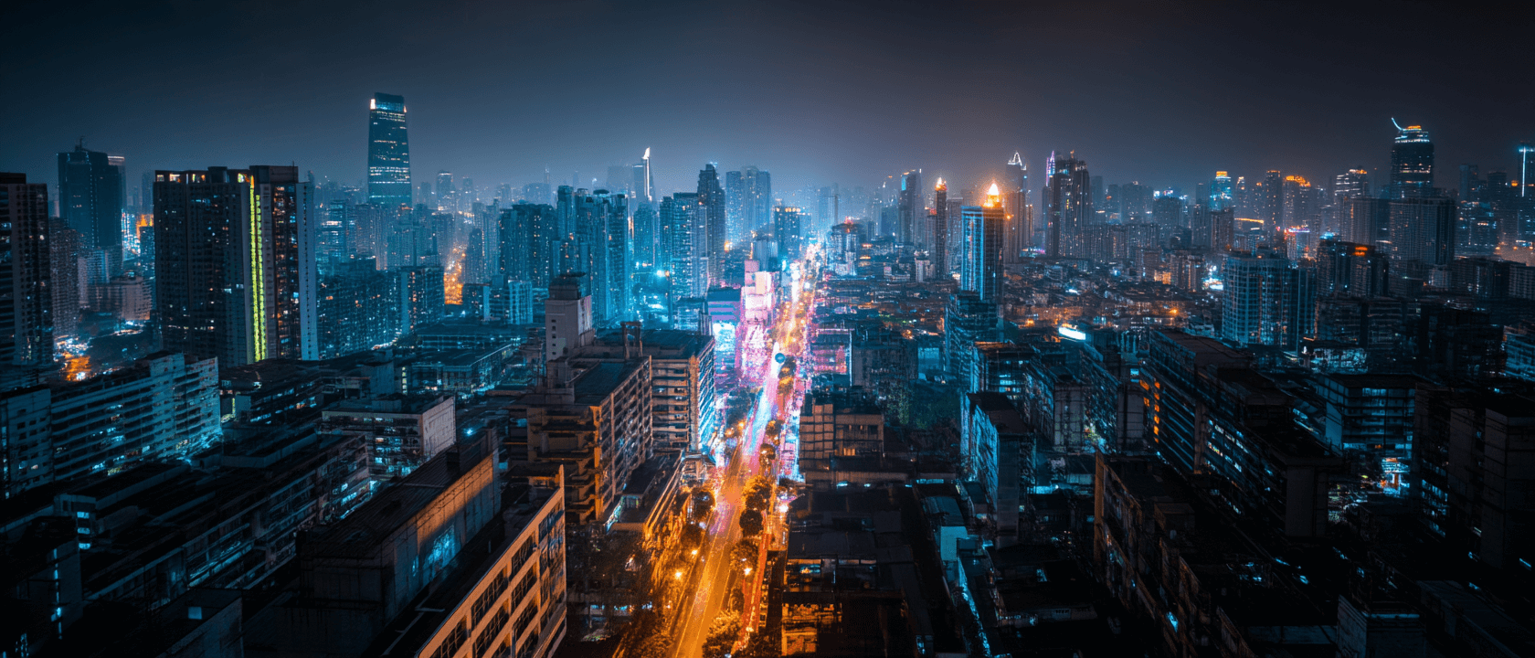 A vibrant cityscape at night with tall skyscrapers and illuminated buildings. Bright neon lights line a busy street running through the center, creating a lively urban atmosphere under a dark, hazy sky.